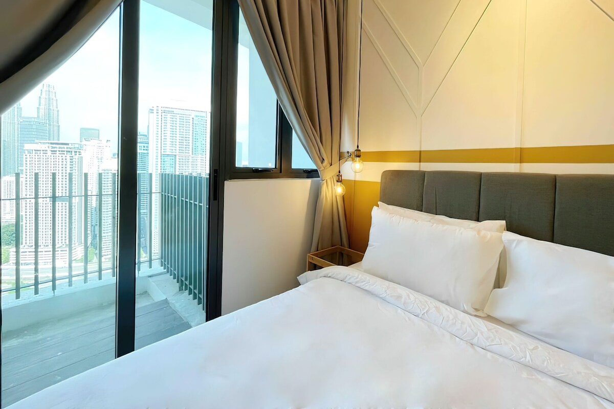 Habitación de hotel con una cama grande, una mesita de noche, una lámpara y vistas de edificios altos a través de un ventanal.