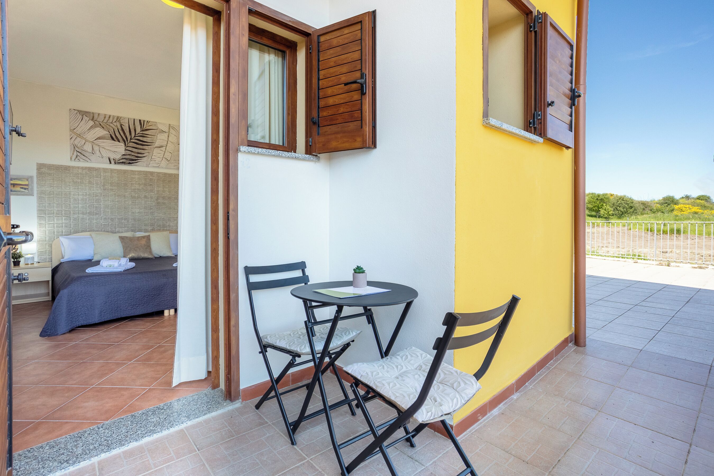 Quarto com cama, uma pequena mesa com cadeiras, janela com persianas e vista para o exterior.