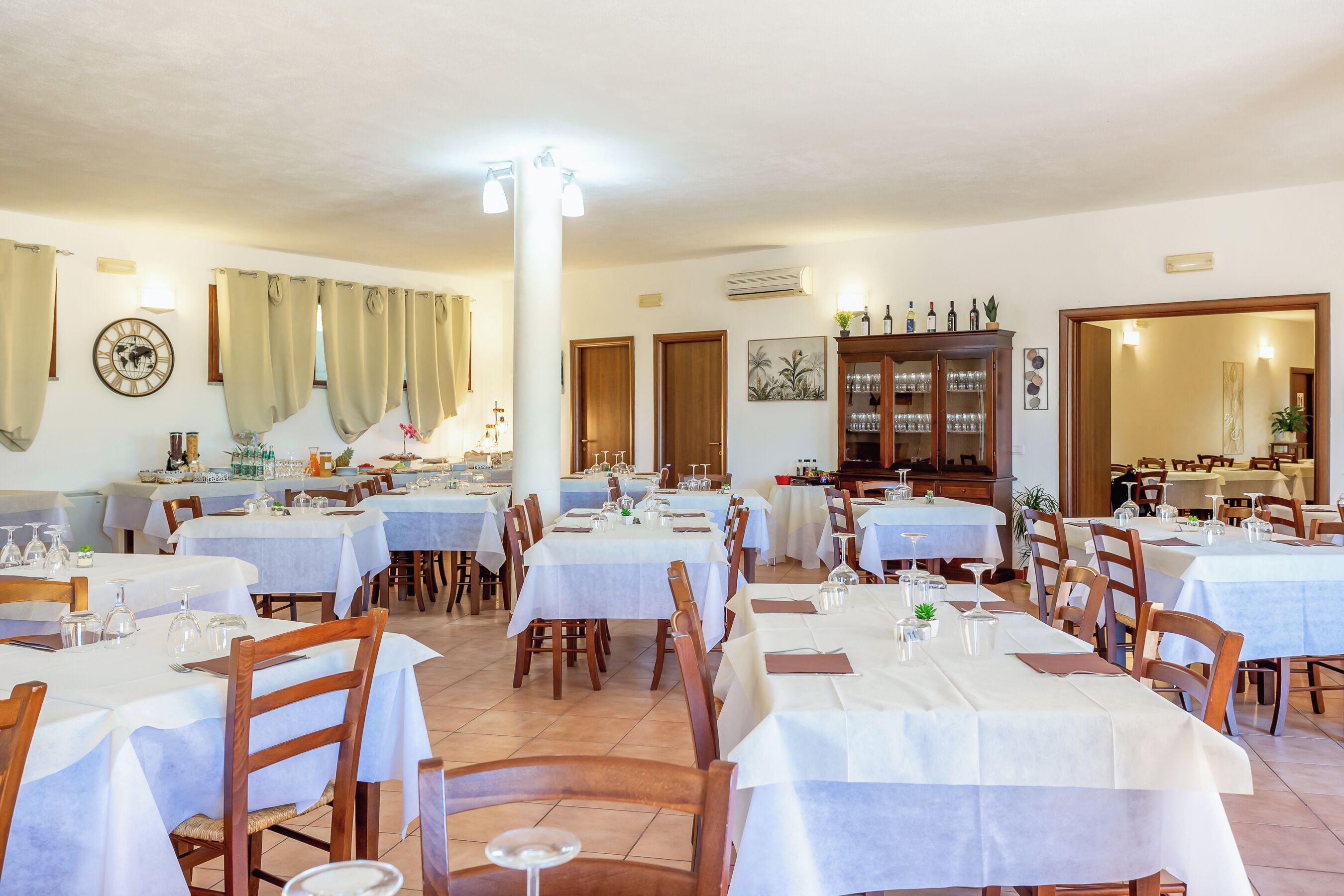 A restaurant with multiple tables and chairs, a clock on the wall, and a cabinet with bottles.