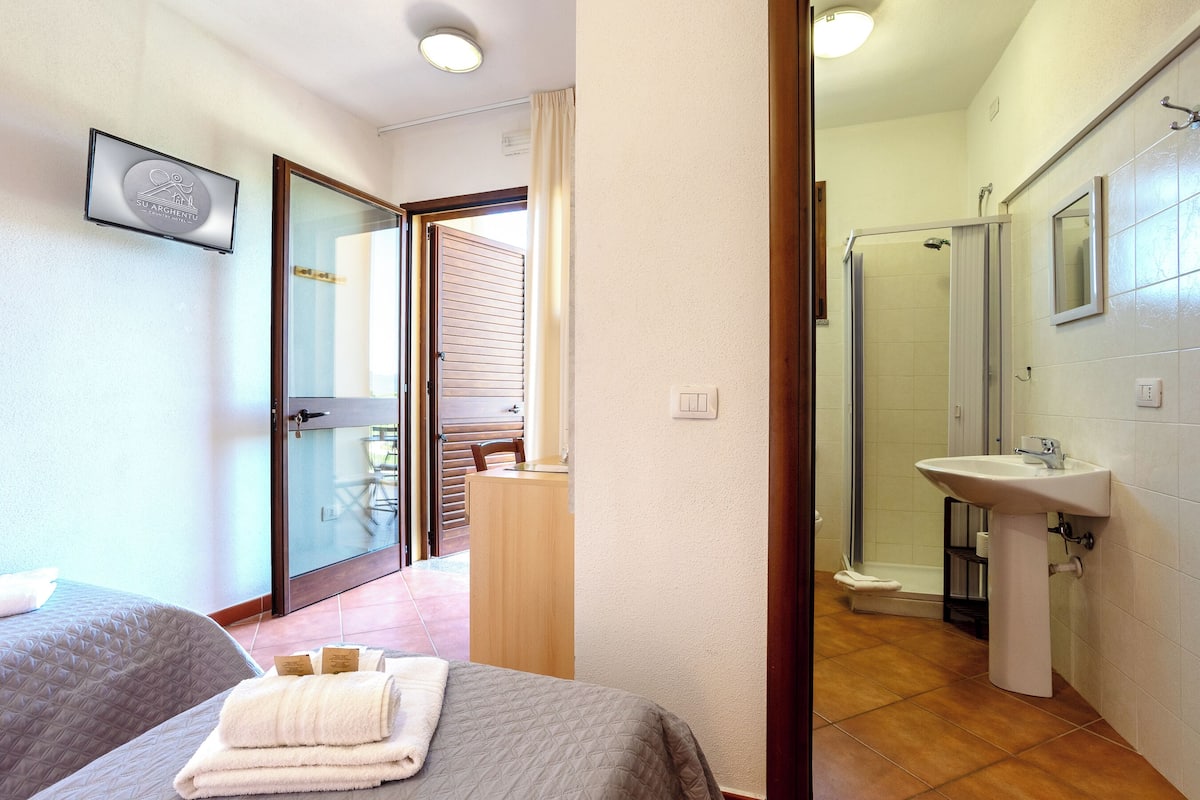 Habitació d'hotel amb llit, taula de nit i bany amb rentapla i dutxa.