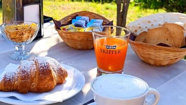 Mesa de comedor al aire libre con croissants, un vaso de jugo de naranja y una taza de café, con árboles y una vista panorámica de fondo.