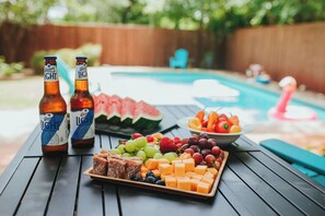 Une table placée au bord de la piscine, avec des bouteilles, une pastèque et un assortiment de fruits et de fromages.