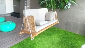 A modern outdoor seating area with a wicker swing chair, green artificial grass, and a wall of various plants. - Riveria City KL Sentral Luxury Apartment (Kuala Lumpur)