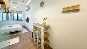 A modern, well-lit room with a bed, a small bar area with stools, a wall-mounted shelf, and a large window offering a city view. - Riveria City KL Sentral Luxury Apartment (Kuala Lumpur)