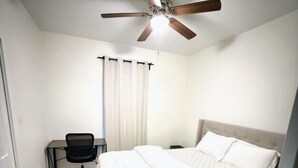 Ein Schlafzimmer mit Bett, Schreibtisch, Stuhl, Deckenventilator und einem Fenster mit Vorhängen.