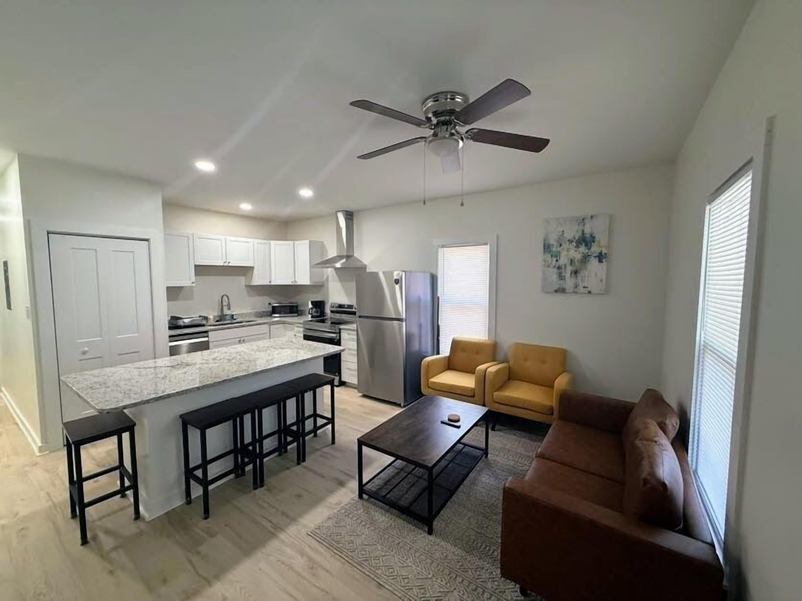 A modern kitchen with white cabinets, a granite countertop, and stainless steel appliances. There is a dining area with a brown sofa and a coffee table.