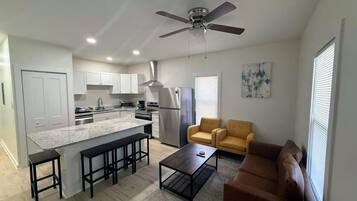 A modern kitchen with white cabinets, a granite countertop, and stainless steel appliances. There is a dining area with a brown sofa and a coffee table.