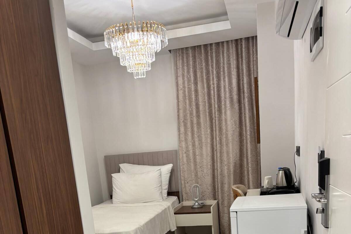 A compact hotel room with a bed, a small table, a refrigerator, and a chandelier.