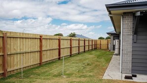 A backyard with a wooden fence, a grassy lawn, and a house with a gray exterior and brick foundation. - Sandy Feet Retreat Su (Peterborough)
