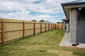 A backyard with a wooden fence, a grassy lawn, and a house with a gray exterior and brick foundation. - Sandy Feet Retreat Su (Peterborough)