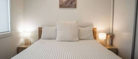 A neatly made bed with striped bedding, two bedside lamps, a framed picture on the wall, and a window on the left.