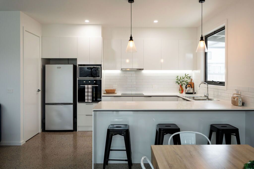 A modern kitchen with white cabinets, a built-in oven, a microwave, and a coffee machine. There are three black stools and a wooden table.