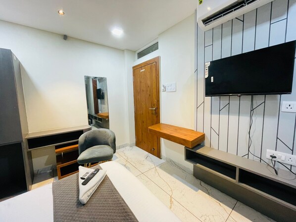Una habitación de hotel moderna con un televisor de pantalla plana montado en la pared, una puerta de madera, una pequeña mesa de madera y un sillón gris.