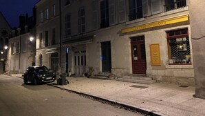 A street view at dusk with a building labeled "LA CURE GOURMANDE" featuring a storefront with a sign and a display window.