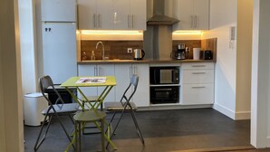 A compact kitchen with white cabinets, a stainless steel range hood, a microwave, and a coffee maker. There is a small dining table with two chairs.