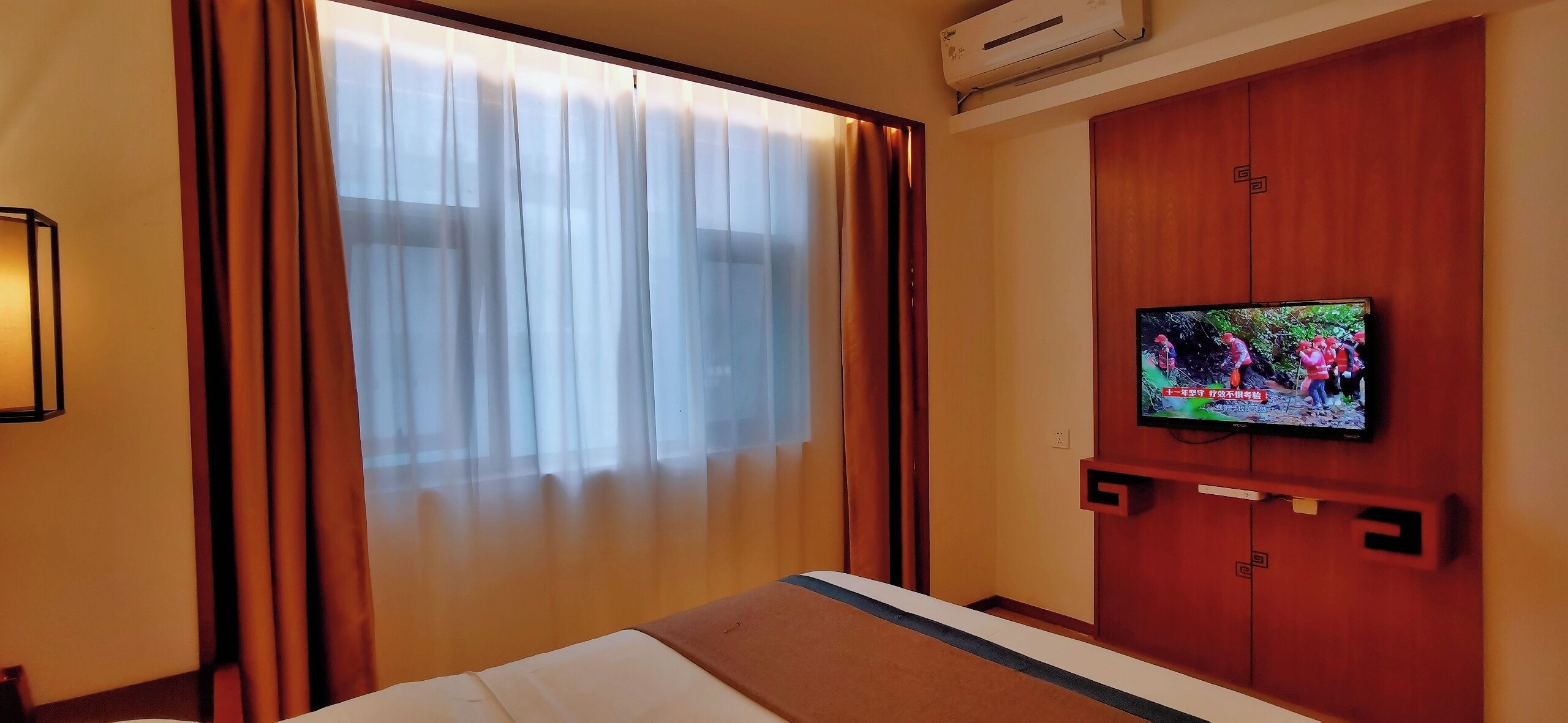 Quarto de hotel com uma cama, uma televisão fixada na parede e uma janela com cortinas.