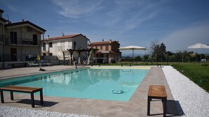 A swimming pool with two benches, surrounded by a pebble border, with buildings in the background. - Casa In Collina (Terre Roveresche)