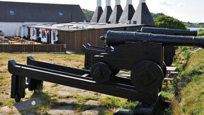 A cannon with a carriage, a wooden fence, and a building with chimneys in the background. - 6 Person Holiday Home in Svaneke (Svaneke)