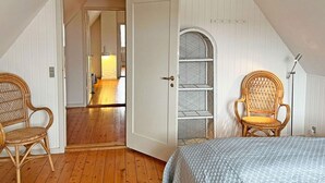 A bedroom with a sloped ceiling, wooden flooring, a bed with a patterned duvet, two wicker chairs, a built-in shelving unit, and a door leading to another room. - 6 Person Holiday Home in Svaneke (Svaneke)