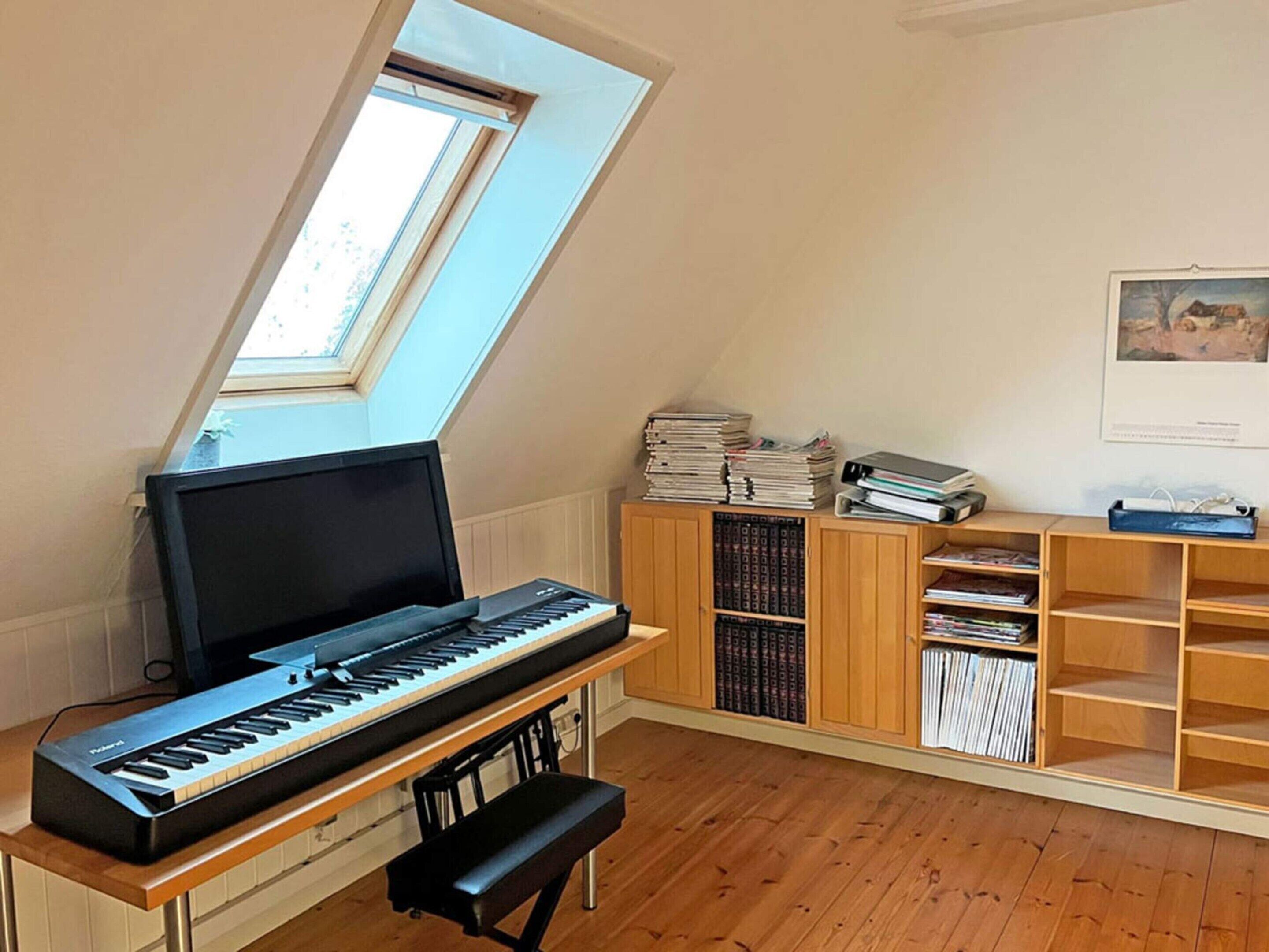 A room with a piano, a bench, a skylight, and a bookshelf filled with books and binders.