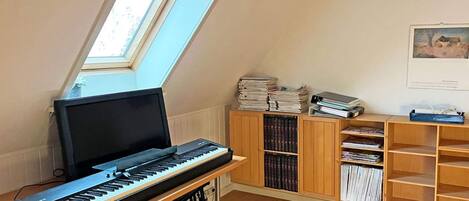 A room with a piano, a bench, a skylight, and a bookshelf filled with books and binders.