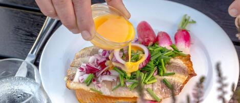 A plate with a fish fillet, radishes, and green onions, with a hand pouring sauce over it.