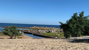A sandy beach area with a rocky shoreline, a small water channel, and a grassy patch with people. - 6 Person Holiday Home in Svaneke (Svaneke)