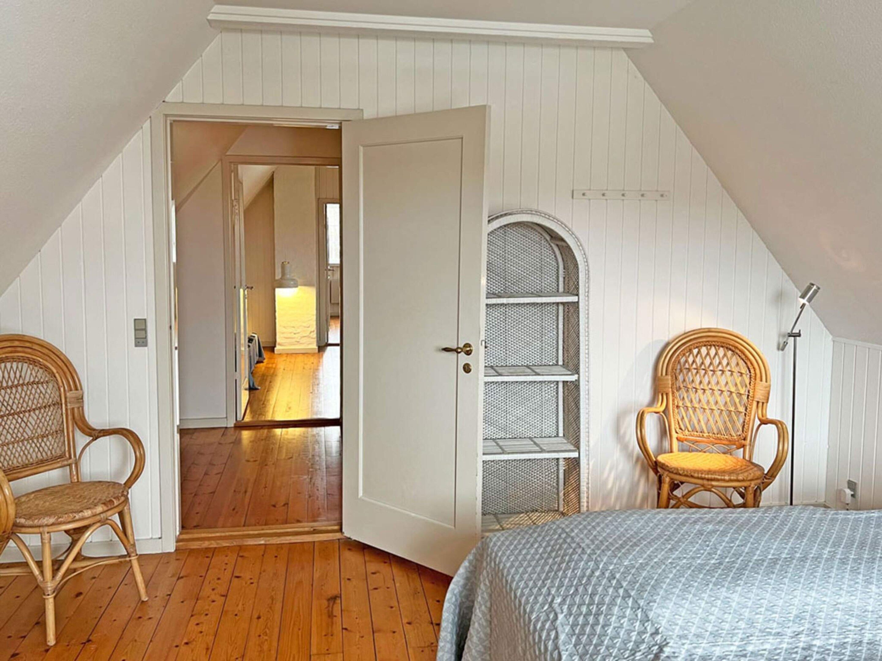 A bedroom with a sloped ceiling, wooden floor, two wicker chairs, a bed with a patterned bedspread, and a built-in shelving unit.
