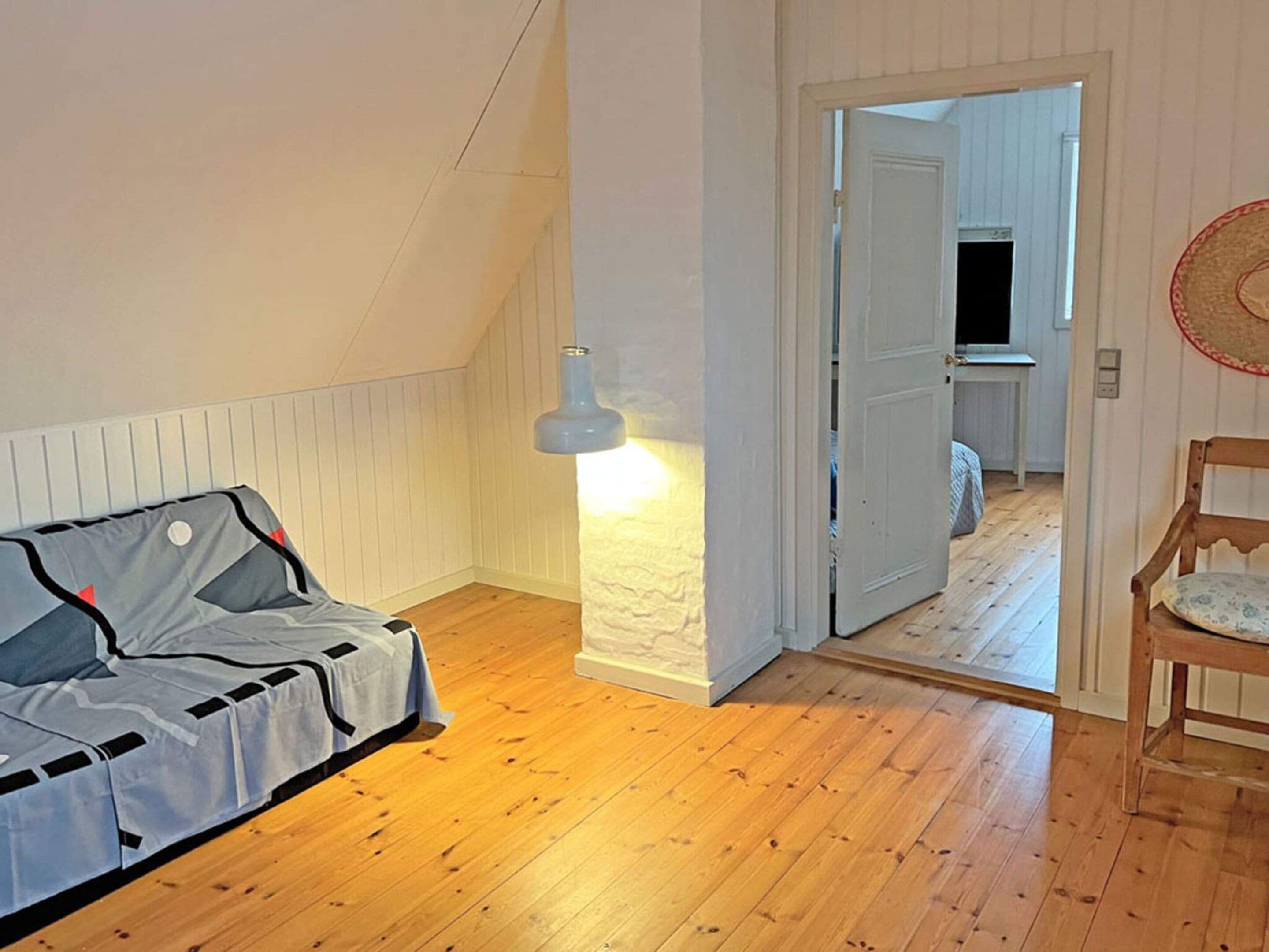 Une pièce avec un plancher en bois, un lit recouvert d’une couverture grise et blanche, un mur blanc avec une partie texturée, et une porte donnant sur une autre pièce.
