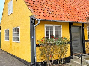 A yellow building with a red-tiled roof, a gray door, and a small garden with red flowers. - 6 Person Holiday Home in Svaneke (Svaneke)