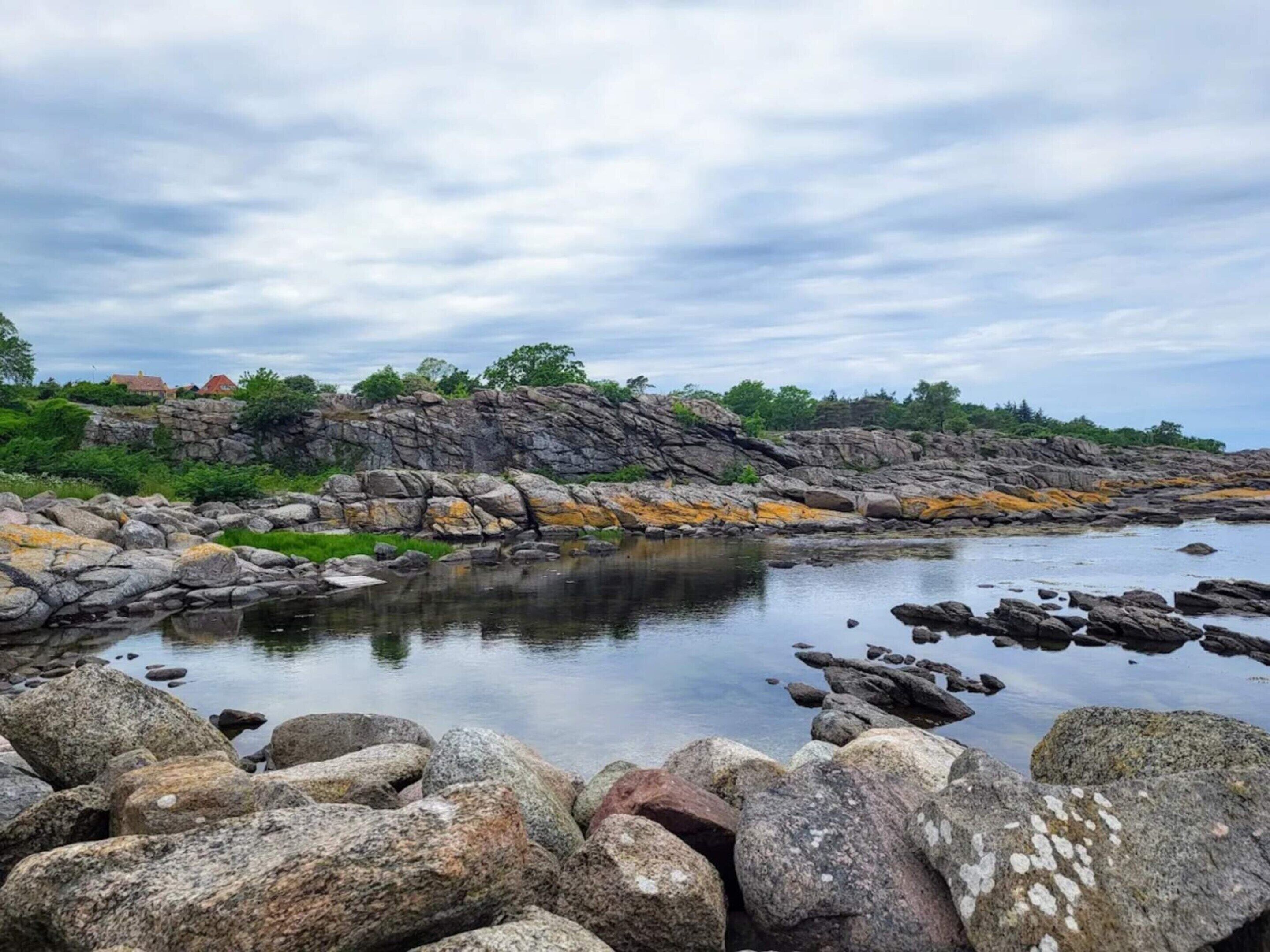 A rocky shoreline with calm water, a stone wall, and greenery in the background.
