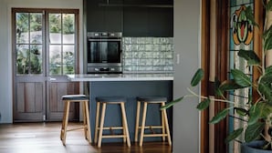 Una cocina moderna con armarios oscuros, horno integrado y un amplio ventanal con vistas a la vegetación. Hay tres taburetes de madera y una planta en maceta.