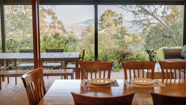 Un espacio de comedor al aire libre con mesas y sillas de madera, y amplios ventanales que ofrecen vistas a un paisaje boscoso.