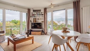 A spacious living room with a large window, a flat-screen TV, a dining area with white chairs, and a bookshelf filled with books and decorative items. - Apartment with port view, parking, near the beach (Hendaye)