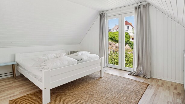 A bedroom with a white bed, a bench, a woven rug, and a view of greenery outside.