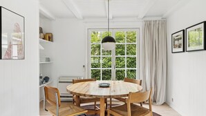 A round wooden table with chairs, a hanging light fixture, a window with curtains, and framed pictures on the wall.