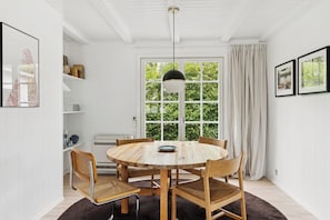 A round wooden table with chairs, a hanging light fixture, a window with curtains, and framed pictures on the wall.