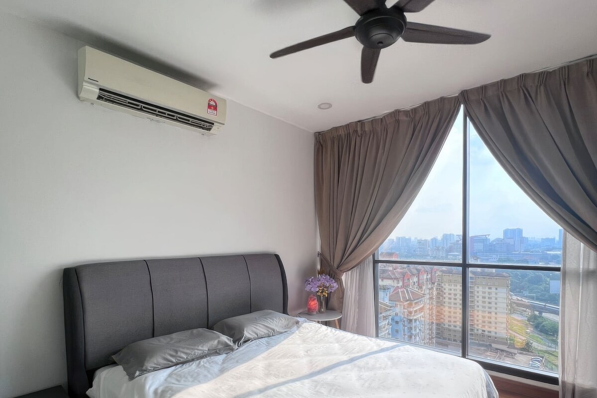 Un dormitorio moderno con una cama grande, ventilador de techo, aire acondicionado y vista a la ciudad a través de amplias ventanas.