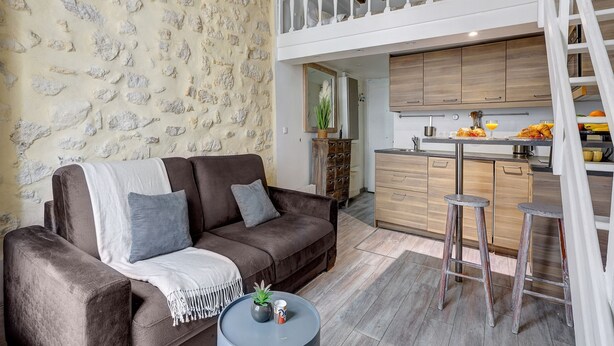 A modern living room with a brown sofa, a coffee table, and a stone wall. A kitchen with wooden cabinets and a balcony is visible in the background.