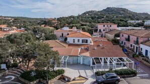 A residential area with terracotta-tiled roofs, a central building with a glass facade, and a hilly backdrop. - Locanda Tartarughino - Suite Luna (Porto Rotondo)