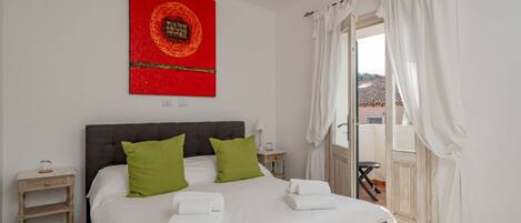 A bedroom with a bed, green pillows, white towels, a nightstand, a red artwork, and a view through a door.