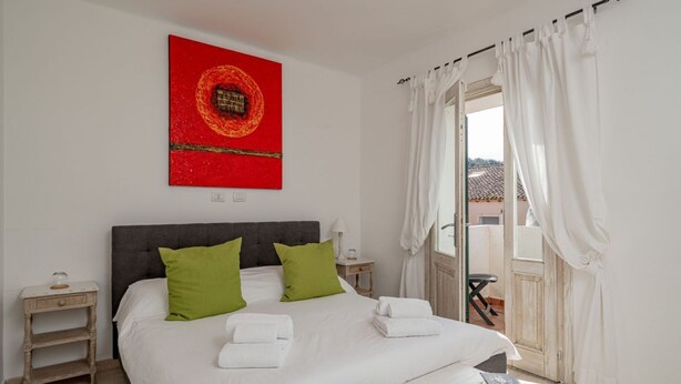 A bedroom with a bed, green pillows, white towels, a nightstand, a red artwork, and a view through a door. - Locanda Tartarughino - Suite Luna (Porto Rotondo)