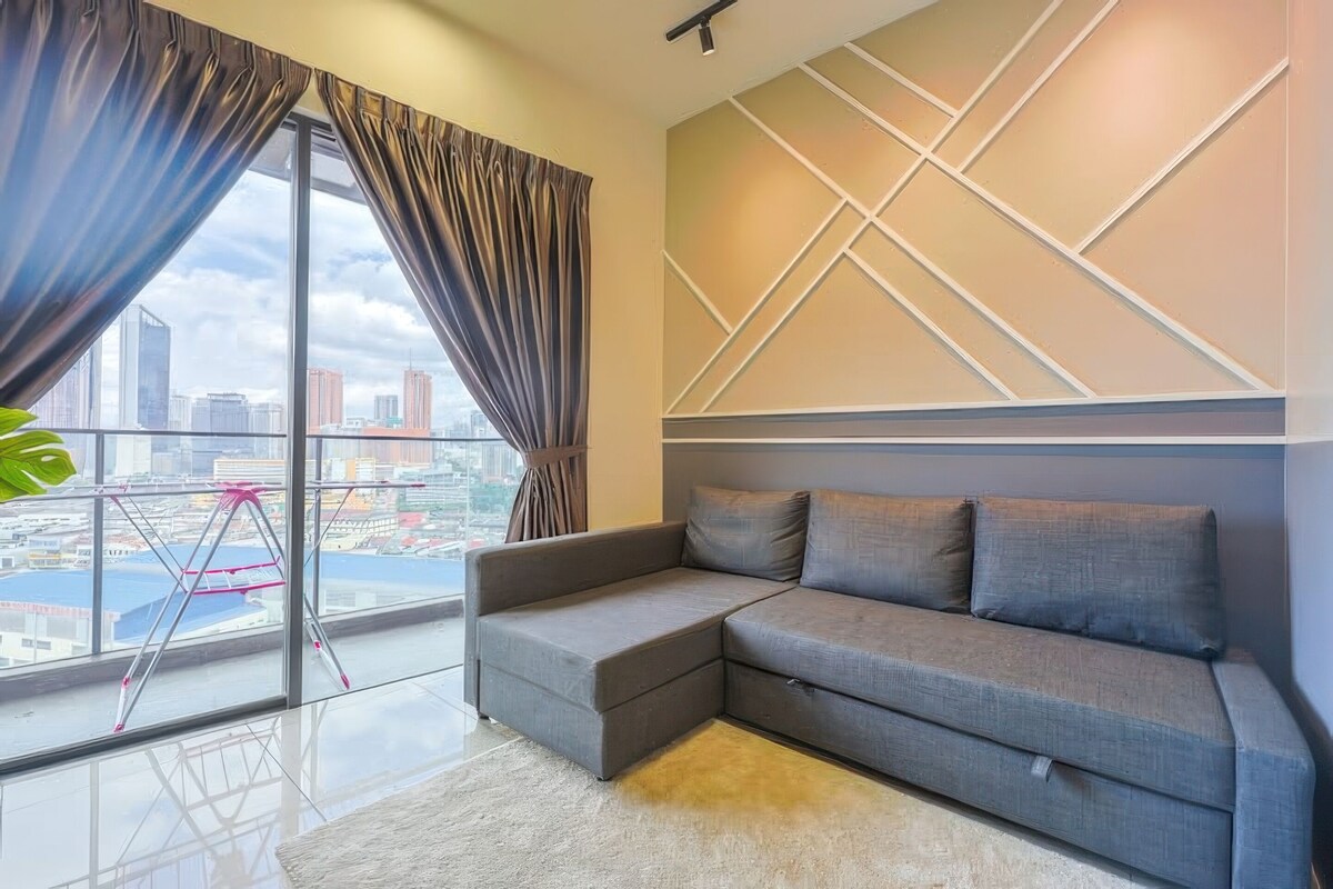 A modern living room with a grey sectional sofa, large windows with curtains, and a city view.