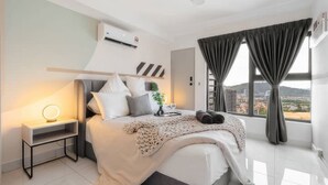 A modern bedroom with a large bed, bedside table with a lamp, and a view of the city through large windows.