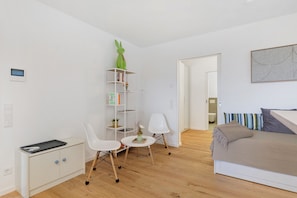 A minimalist bedroom with a bed, a white desk with a computer, a chair, a small table with a vase, and a bookshelf.