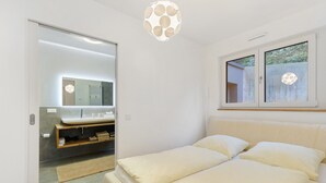 Een slaapkamer met een bed, een raam, een spiegel en een badkamer met een wastafel en handdoekhouder.