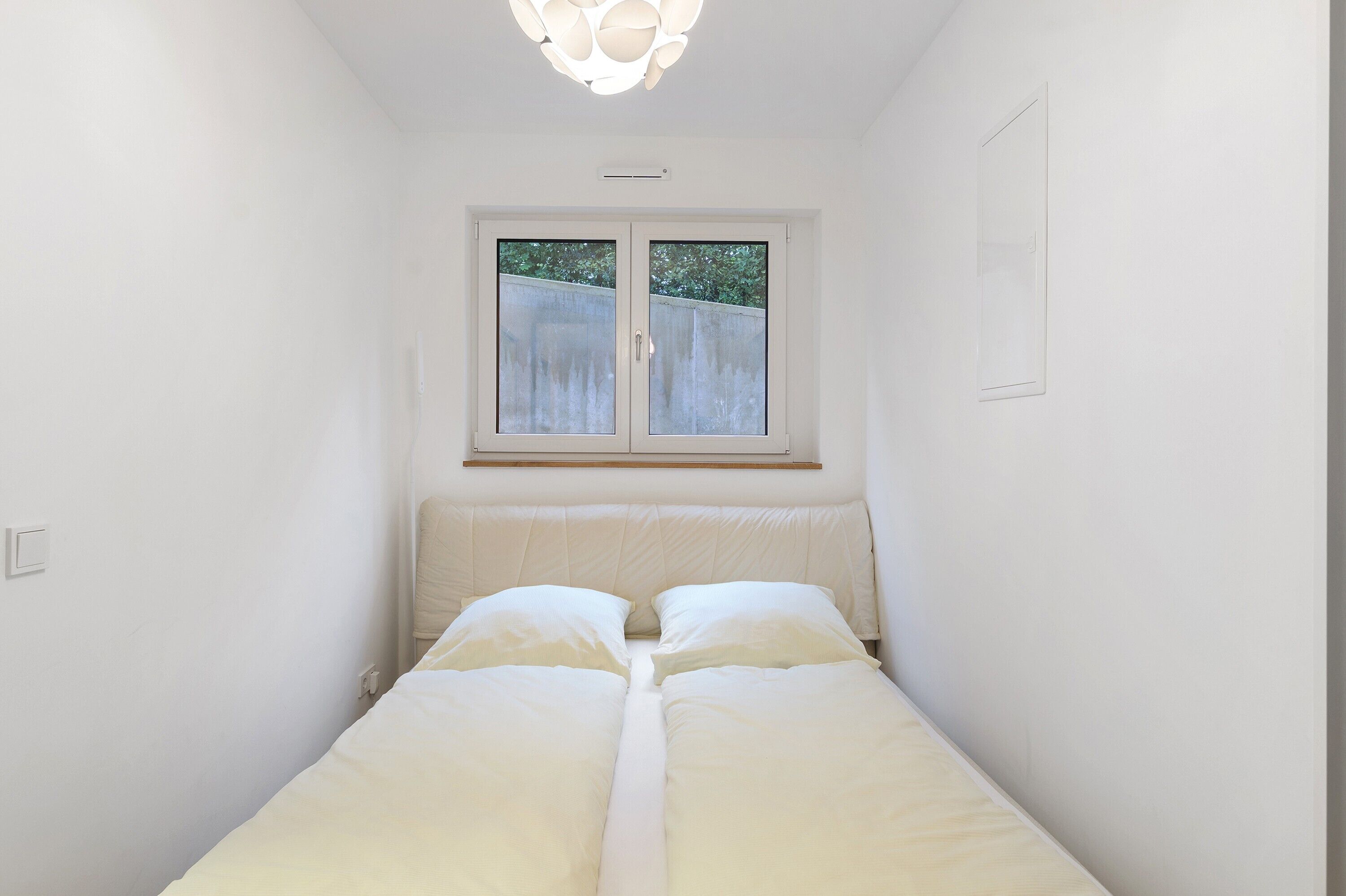 A small, white room with a bed, a window, and a light fixture.