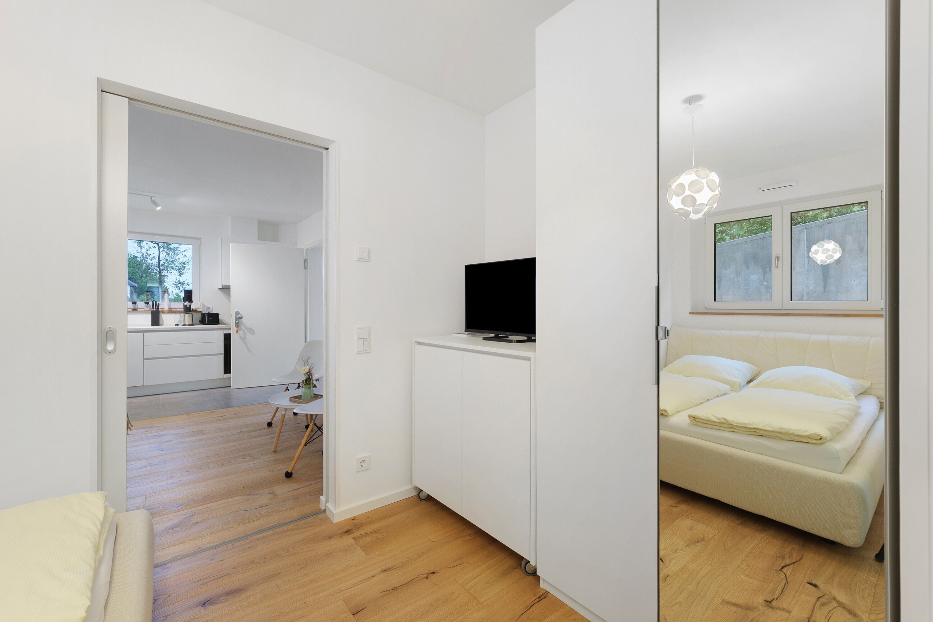 A modern room with a bed, a TV on a white cabinet, and a view of the outside through a window.