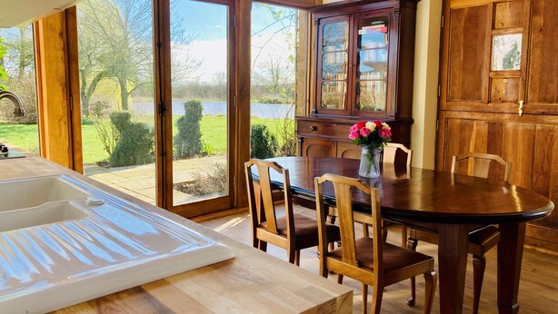 A dining area with a wooden table and chairs, a vase with flowers, and a view of a lake through large glass doors.