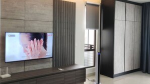 Una sala de estar moderna con un televisor de pantalla plana montado en una pared de paneles de madera, un equipo de aire acondicionado integrado, un mueble de color oscuro y un ventanal con cortinas.